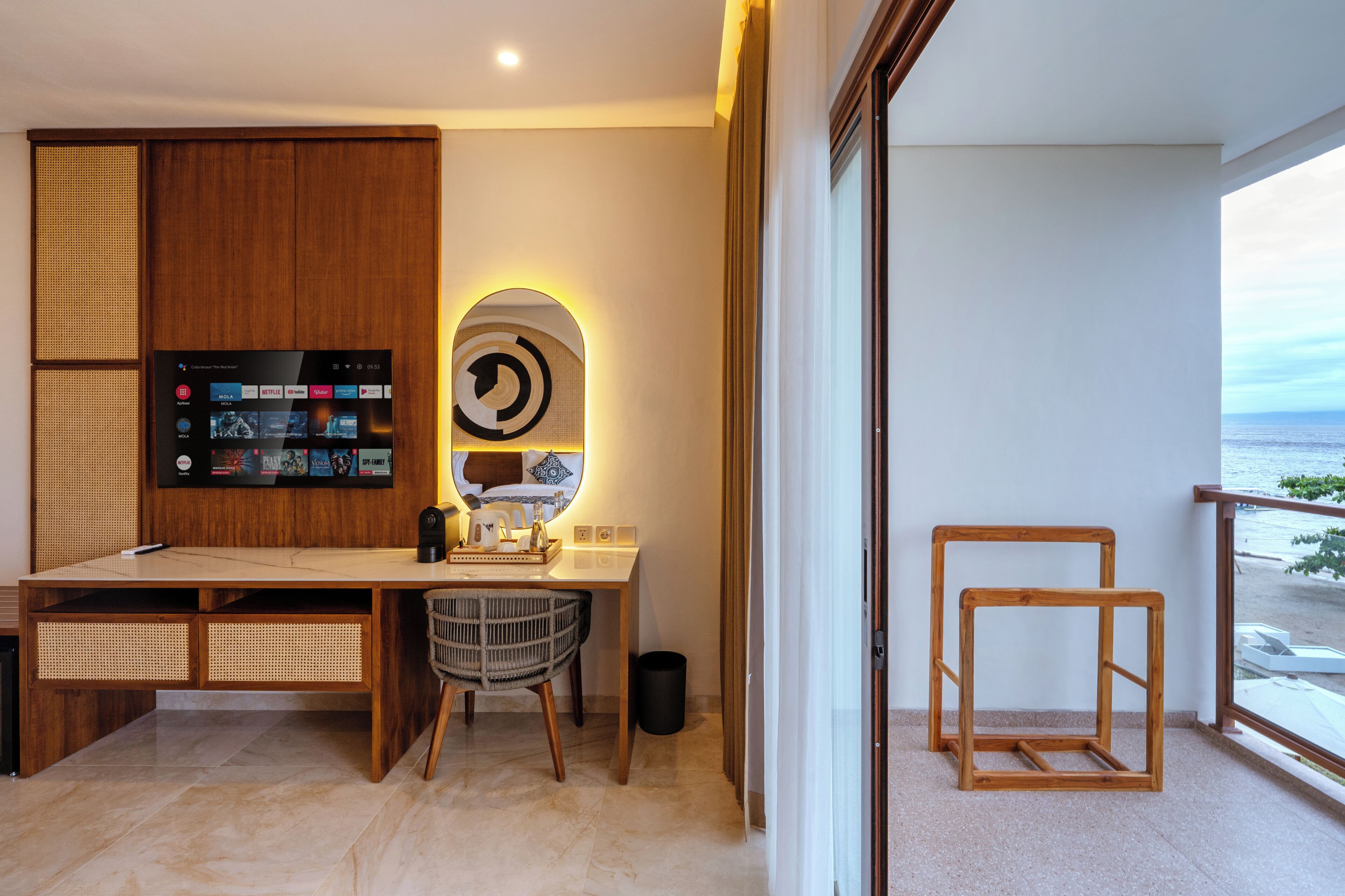 A modern room with a wooden desk, a chair, a TV mounted on the wall, and a balcony with a view of the beach.