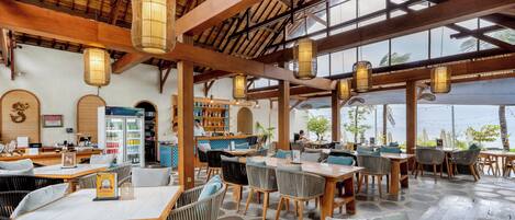 A spacious restaurant with wooden tables and chairs, hanging lanterns, and a view of the sea.