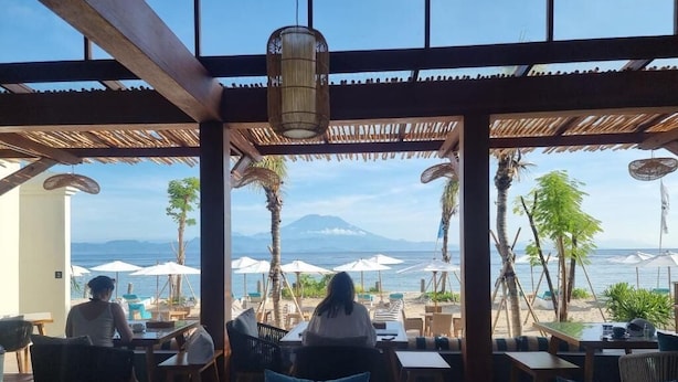 Ein Außenbereich mit Tischen und Stühlen, der auf einen Strand mit Sonnenschirmen und Palmen blickt.