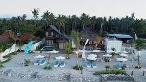 Ein Resort mit mehreren Gebäuden, einem Strandbereich mit Liegestühlen und Sonnenschirmen und einer Kulisse aus Palmen.