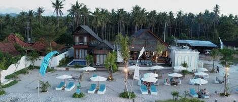 A resort with multiple buildings, a beach area with lounge chairs and umbrellas, and a backdrop of palm trees.