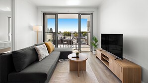 Een moderne woonkamer met een grijze bank, een houten salontafel, een televisie op een houten kast en een balkon met tuinmeubilair.
