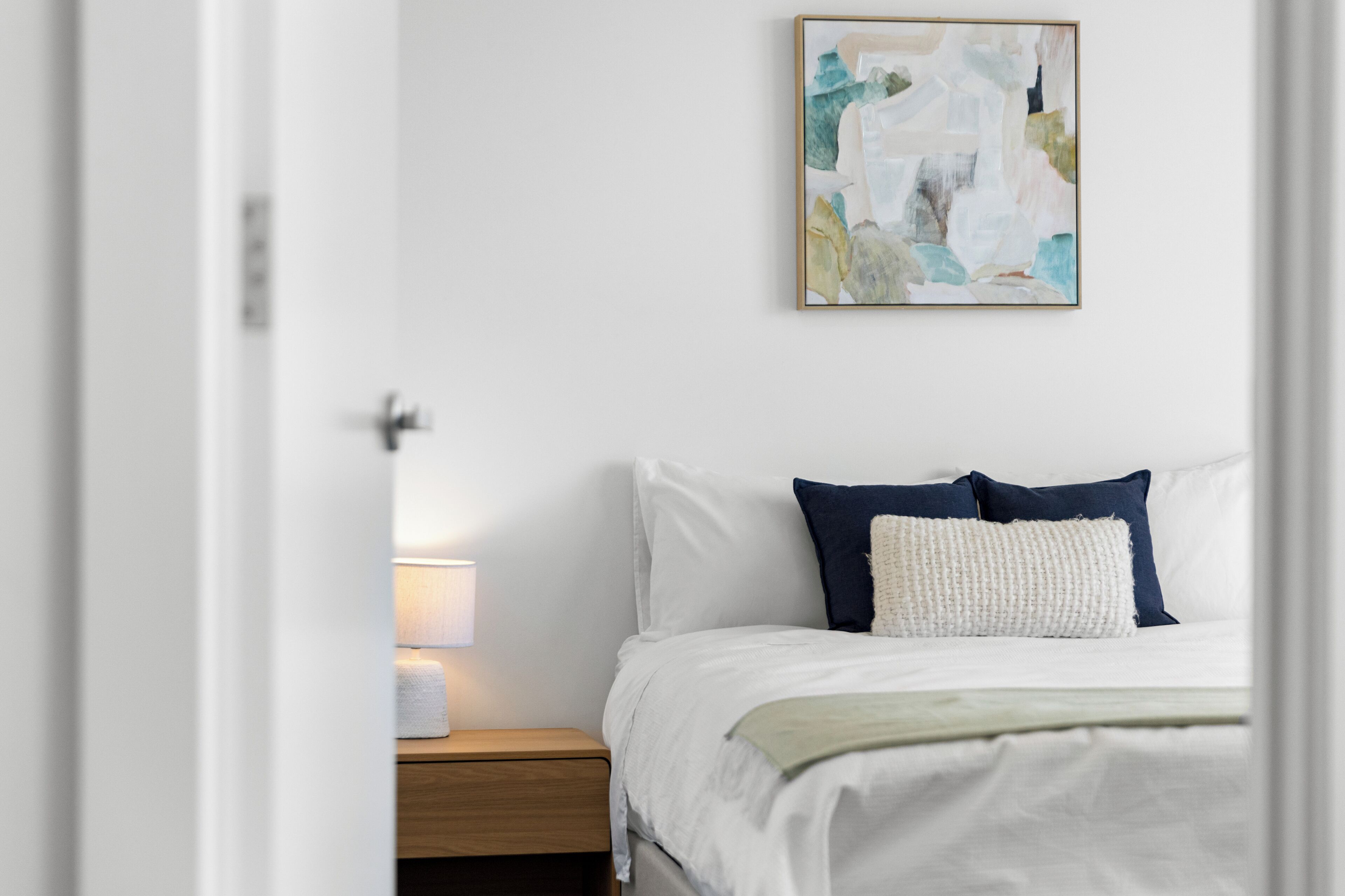 A bedroom with a bed, pillows, a nightstand, a lamp, and a painting on the wall.