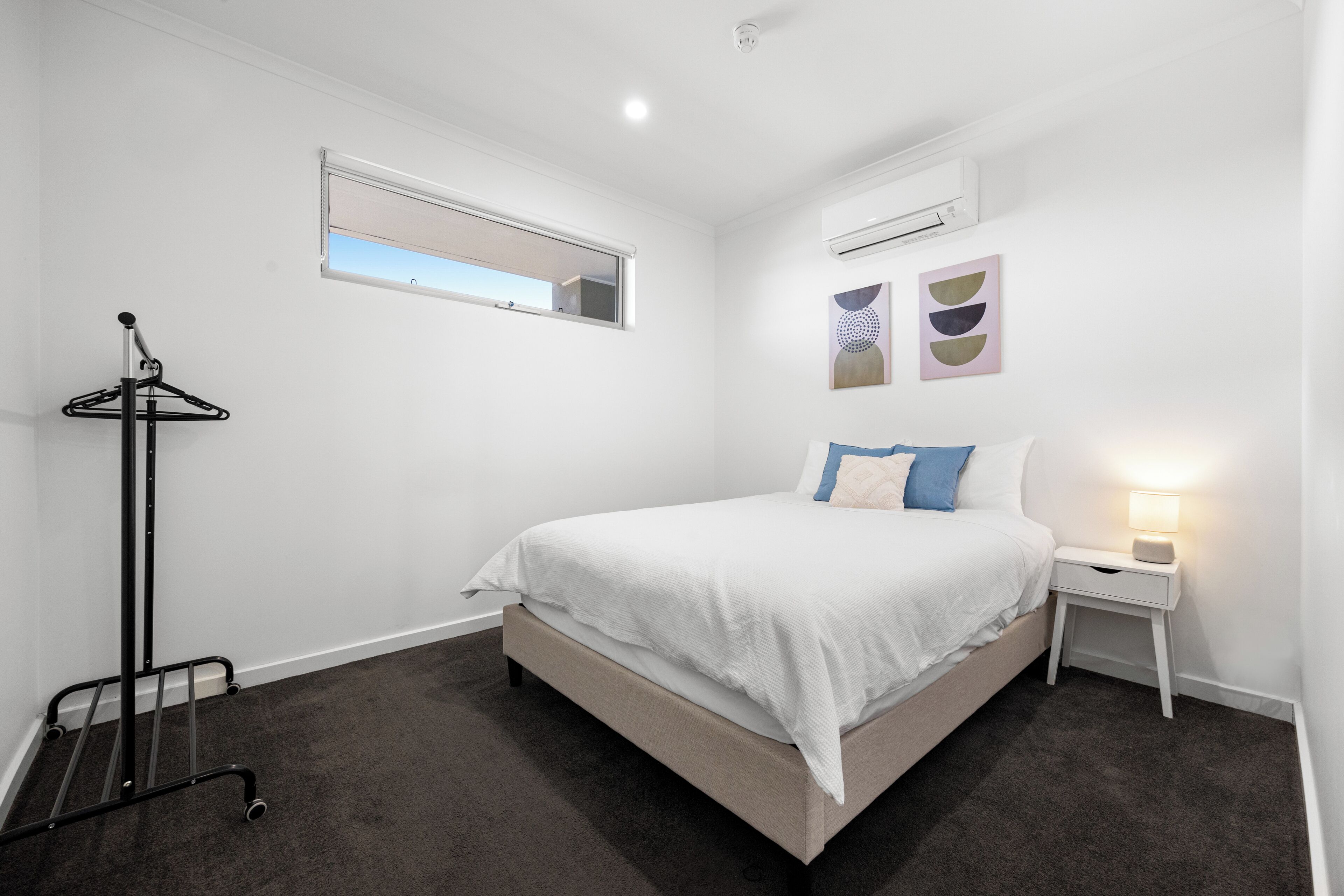 Un dormitorio con cama, mesita de noche con lámpara, perchero, ventana y decoración en la pared.