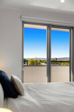 A bedroom with a bed, pillows, and a large window offering a view of a balcony and distant mountains. - Contemporary 2BR Apartment | Heart of Prospect (Prospect)