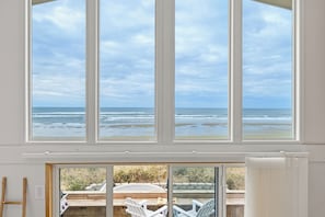 Ein Zimmer mit großen Fenstern mit Blick aufs Meer, eine Holzterrasse mit Stühlen und eine Aussicht auf den Strand.