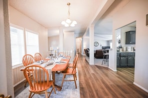 A dining area with a wooden table and chairs, a chandelier, and a kitchen in the background.