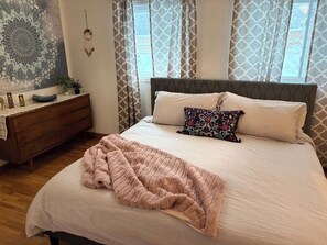 Ein ordentlich bezogenes Bett mit gemustertem Überwurf, eine Holzkommode mit Spiegel und eine Wandteppich.