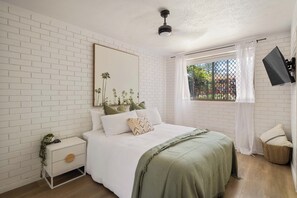 Een slaapkamer met een witte bakstenen muur, een bed met een groene deken, een nachtkastje met een plant en een raam met uitzicht op het omliggende landschap.