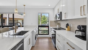 Une cuisine moderne avec des meubles blancs, des appareils en acier inoxydable et une grande fenêtre donnant sur un quartier résidentiel.