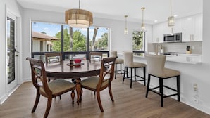 Une cuisine moderne avec des meubles blancs, un coin repas avec des chaises en bois et une grande fenêtre donnant sur l’extérieur.