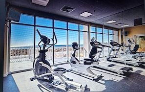 A gym with various exercise machines, large windows, and a view of the beach. - Coastal Breeze Retreat (Navarre Beach)