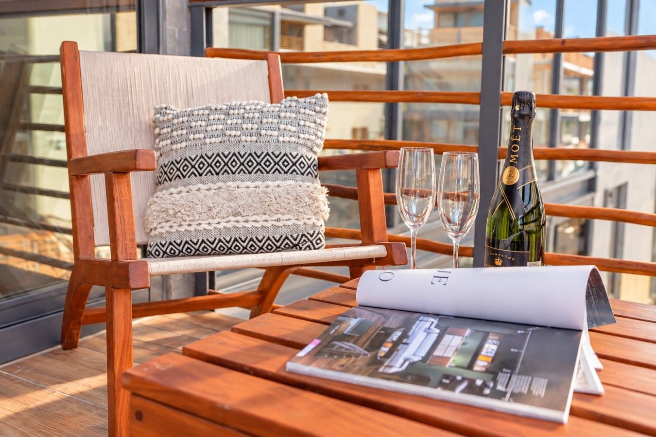 A wooden chair with a patterned cushion, two wine glasses, a bottle of champagne, and a menu on a wooden table.