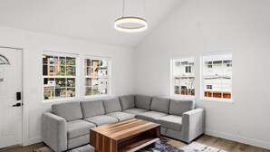 Un salon moderne avec un canapé d’angle, une table basse en bois et de grandes fenêtres donnant sur des immeubles et des arbres.