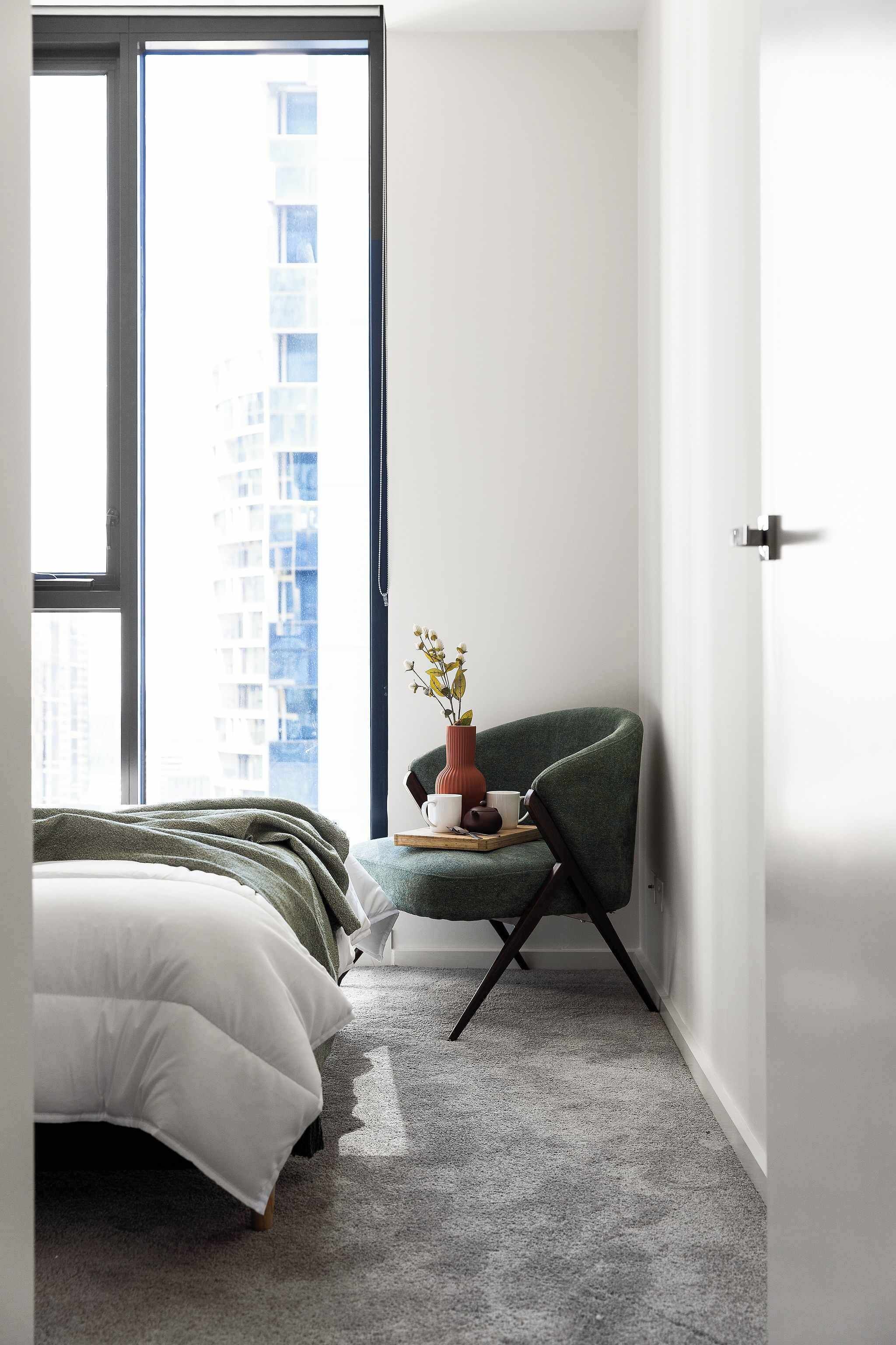 Ein Schlafzimmer mit einem Bett, einem Sessel, einem kleinen Tisch mit einer Vase und Blick auf ein Gebäude durch ein Fenster.