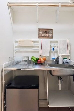 A compact kitchenette with a sink, a small dish rack, a bowl of fruit, and a coffee sign on the wall.