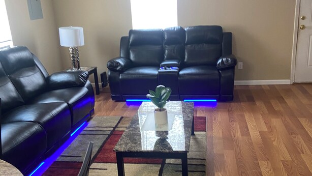 A living room with a black leather sofa, a glass coffee table with a plant, and a ceiling fan.