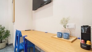 Une petite pièce, bien rangée, avec une table en bois, des chaises bleues, un téléviseur fixé au mur, une petite plante et une machine à café Krups.