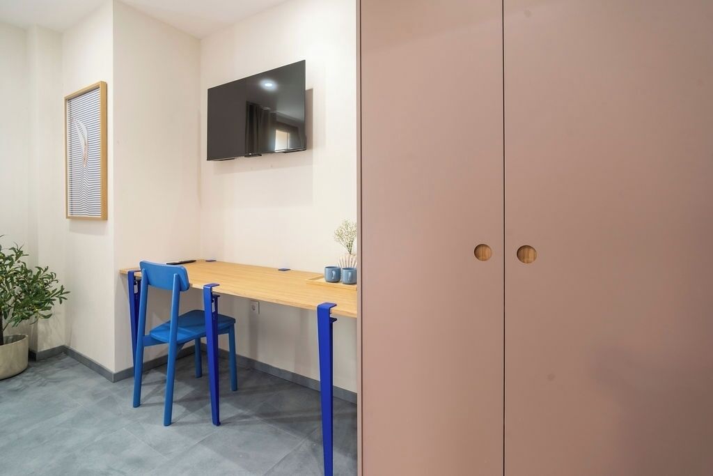 Una habitación con una mesa de madera, dos sillas azules, una planta en maceta, un televisor montado en la pared y un mueble rosa grande.