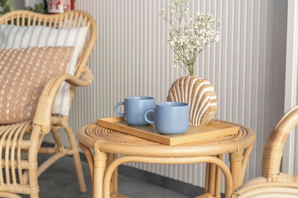 Una silla de mimbre con cojín, una mesita redonda pequeña con una bandeja que contiene dos tazas azules y un florero de concha decorativo con flores.