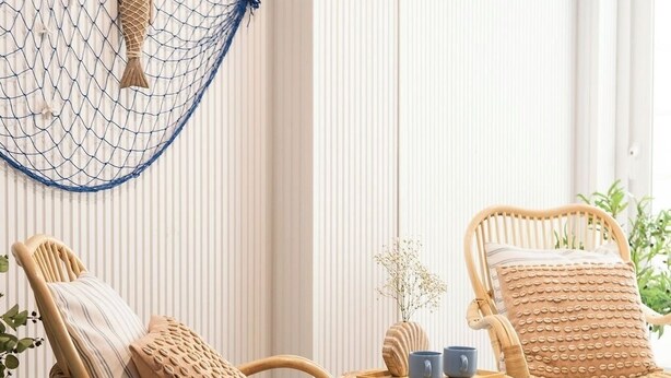 Twee rieten stoelen met kussens, een klein tafeltje met twee blauwe mokken, en een decoratieve vis die aan een net aan de muur hangt.