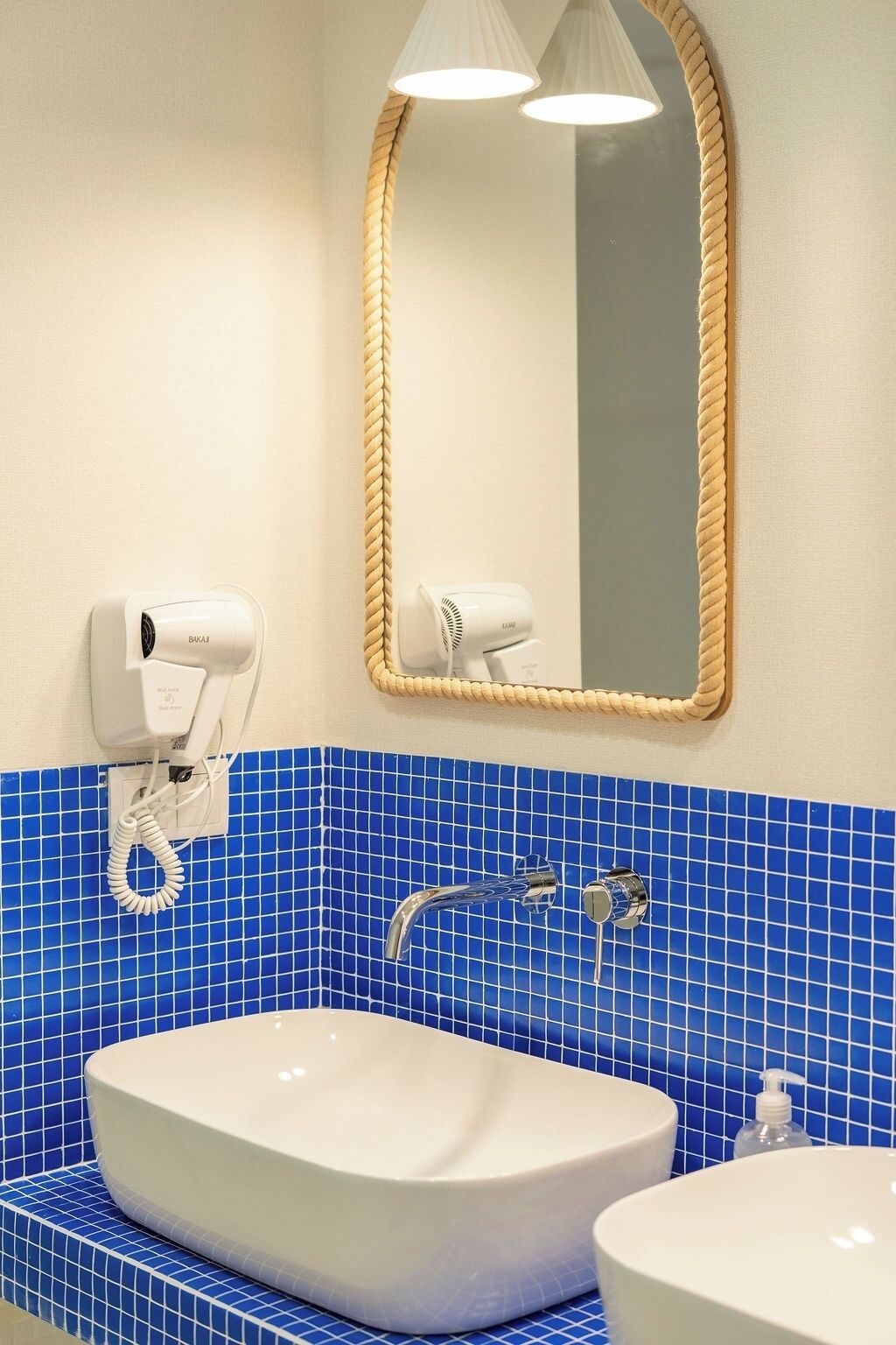 Un baño con lavamanos blanco, salpicadero de azulejos azules, un espejo redondo con marco de cuerda y dos secadores de pelo montados en la pared.