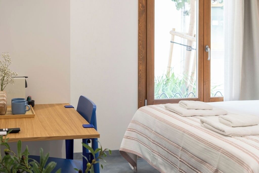 A hotel room with a bed, a wooden desk with a chair, a small table with a vase and a tray, and a window with a view of greenery.