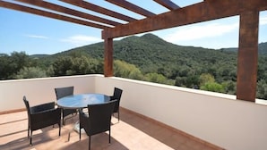 Ein Balkon mit rundem Tisch und Stühlen, Blick auf eine bewaldete Bergkette und einen klaren Himmel.