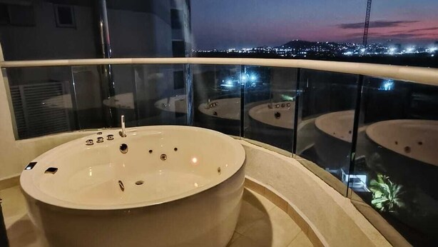A modern bathroom with a large bathtub, a glass-enclosed balcony with a city view at dusk, and a curved floor design. - Oceani Cartagena (La Boquilla)