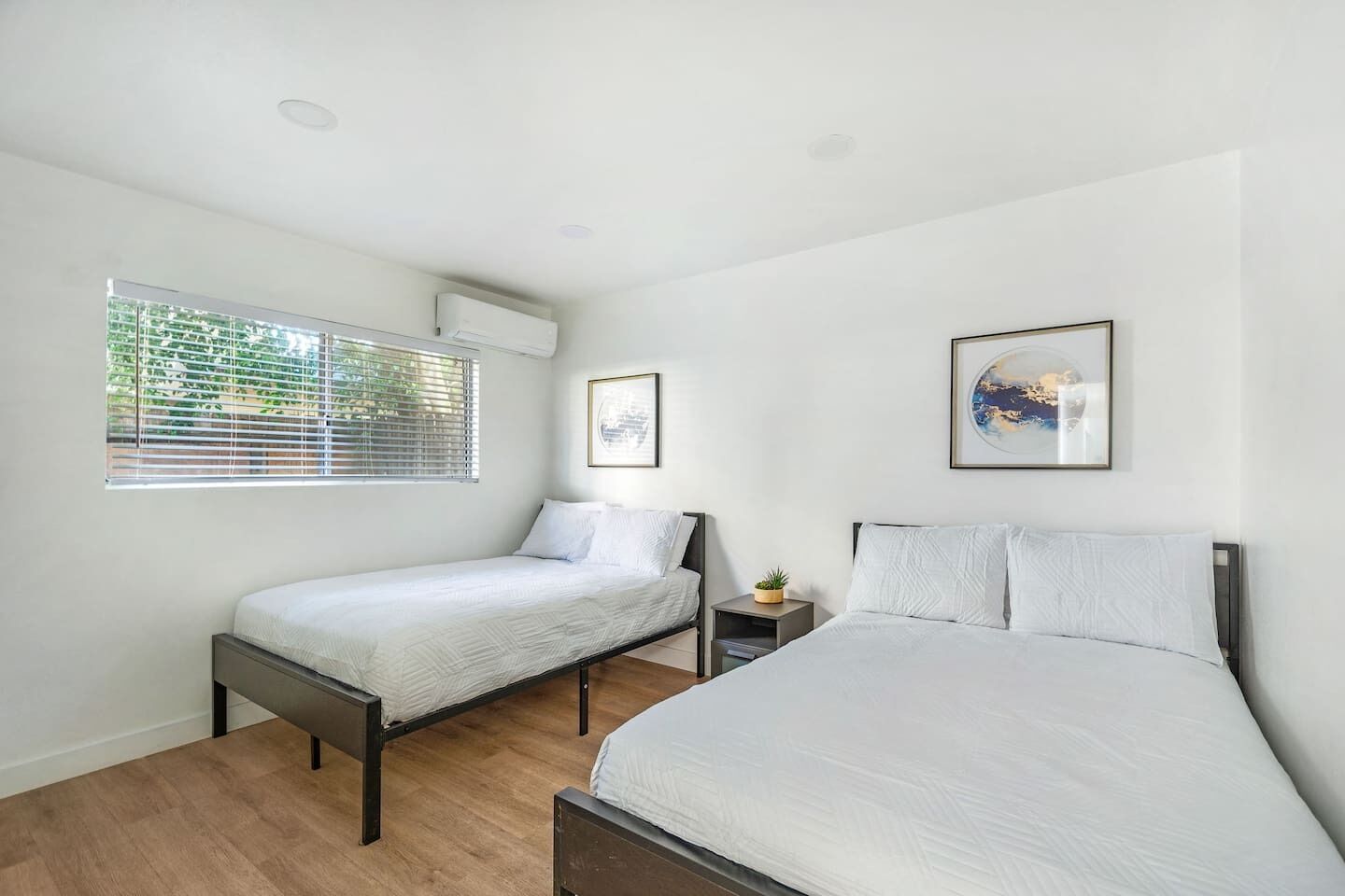 Een slaapkamer met twee eenpersoonsbedden, een raam met zonwering, een airconditioningapparaat en twee ingelijste foto's aan de muur.