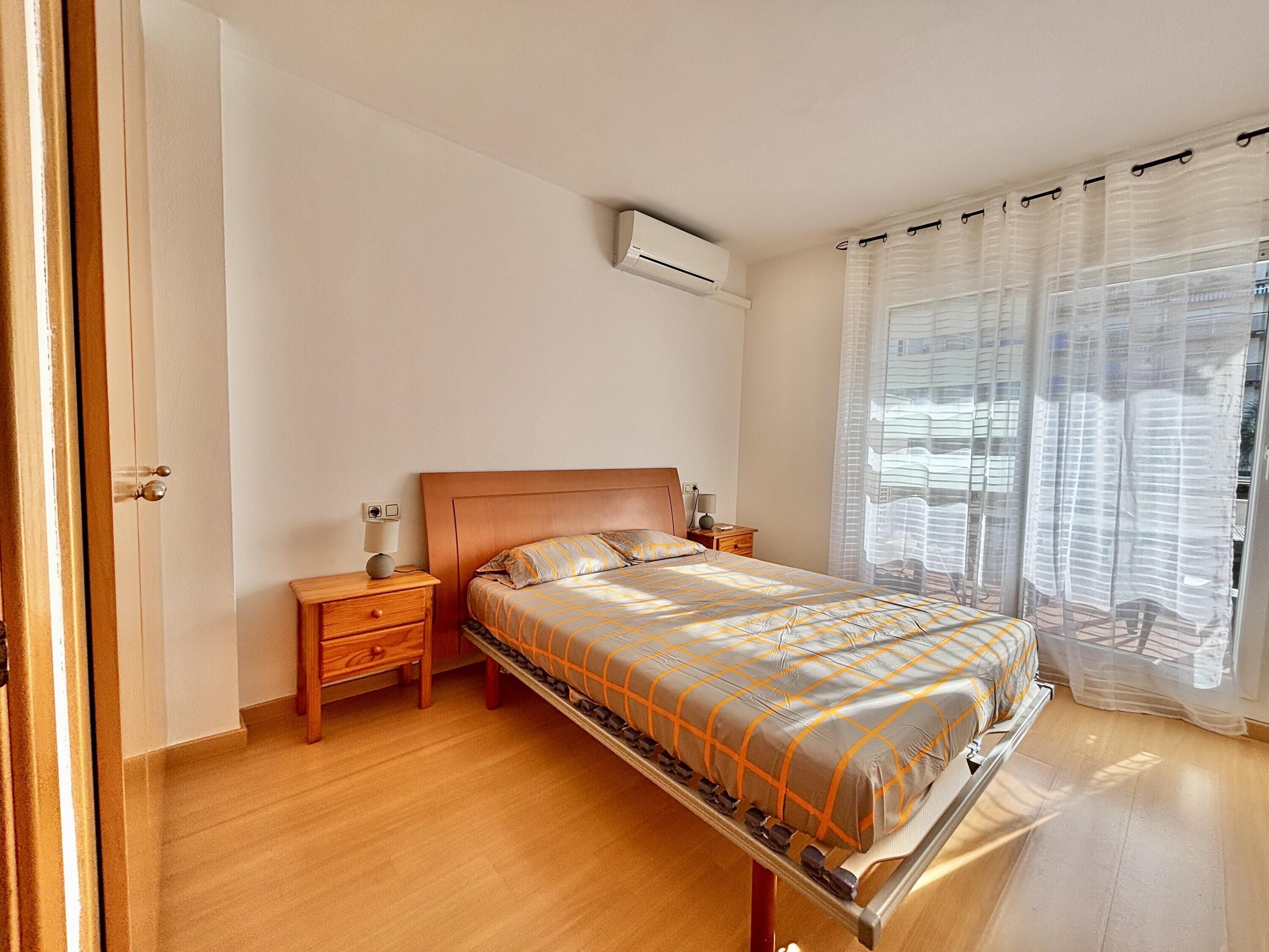 Une chambre à coucher avec un lit en bois, une table de chevet, un climatiseur fixé au mur et des rideaux légers.