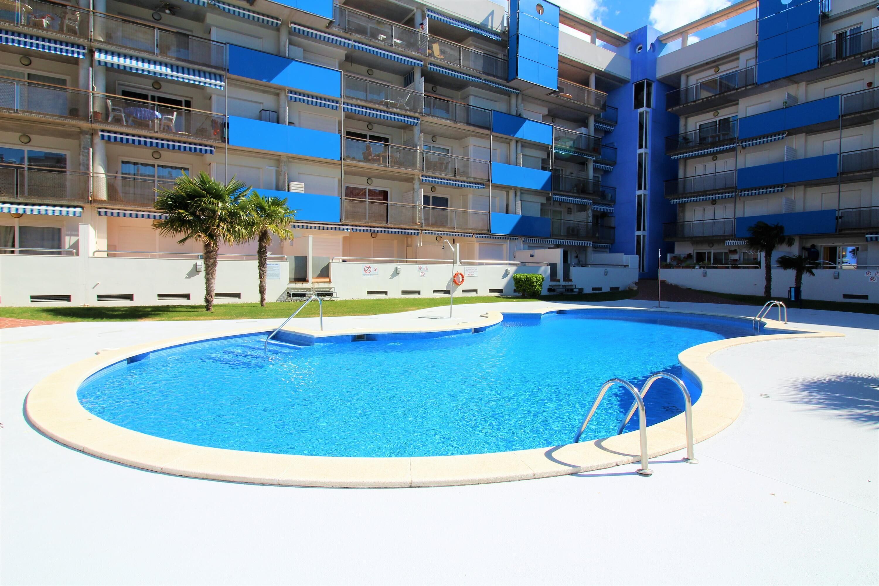 A swimming pool with a circular shape, surrounded by a white deck. In the background, there is a multi-story building with balconies and a blue facade.