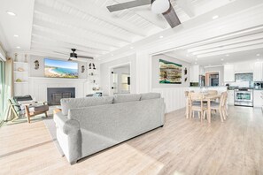 A spacious living room with a grey sofa, wooden dining table, and chairs. A fireplace is visible in the background, and there is a kitchen area with modern appliances. - 3BR Beachfront Home, Patio on the Sand w/ Gorgeous Ocean Views! (Newport Beach)