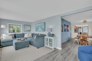 A living room with a sectional sofa, a coffee table, a cabinet with magazines, and a dining area with a table and chairs. - Lakefront Cottage, Dock, EV Charger, Firepit, Bunkhouse, Family & Pet Friendly (Carp Lake)
