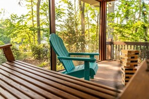 A wooden deck with a green Adirondack chair and a table, surrounded by a forest view. - Lake Sinclair Waterfront Getaway | Private Dock | Screened Porch (Eatonton)