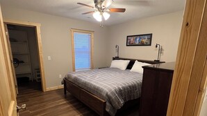 Une chambre à coucher avec un lit, un plancher en bois, un ventilateur de plafond, une fenêtre avec des stores et un tableau au mur.