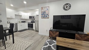 Une cuisine moderne avec des meubles blancs, un four à micro-ondes et un réfrigérateur. On y trouve un coin repas avec une table et des chaises noires, ainsi qu’un grand téléviseur à écran plat posé sur un meuble en bois.
