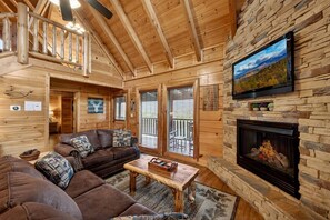 Ein gemütliches Blockhaus-Interieur mit Holzbalkendecke, Kamin, einer Couch und einem an einer Steinwand befestigten Fernseher.