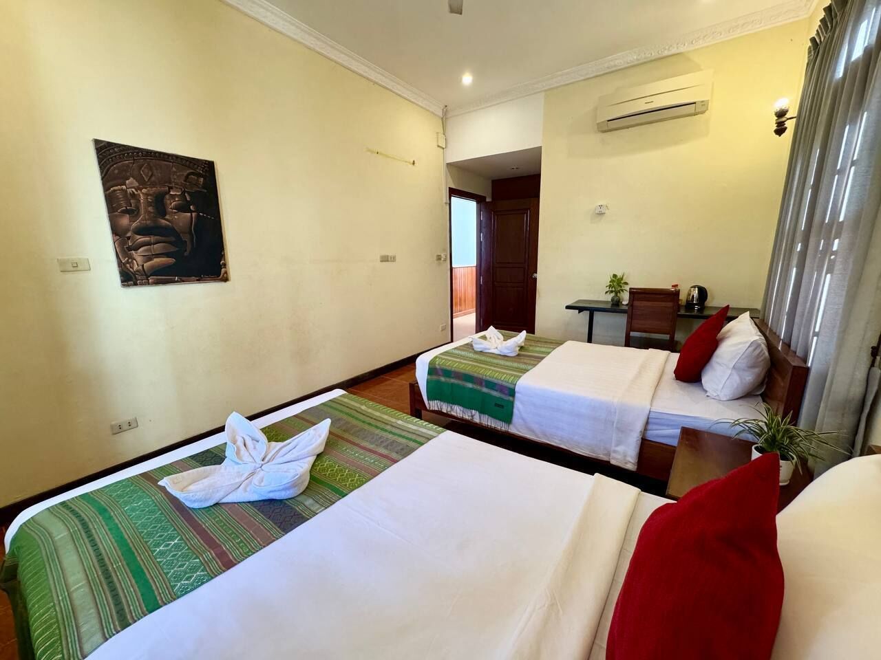 A hotel room with two beds, a small table, a chair, a potted plant, and a painting on the wall.