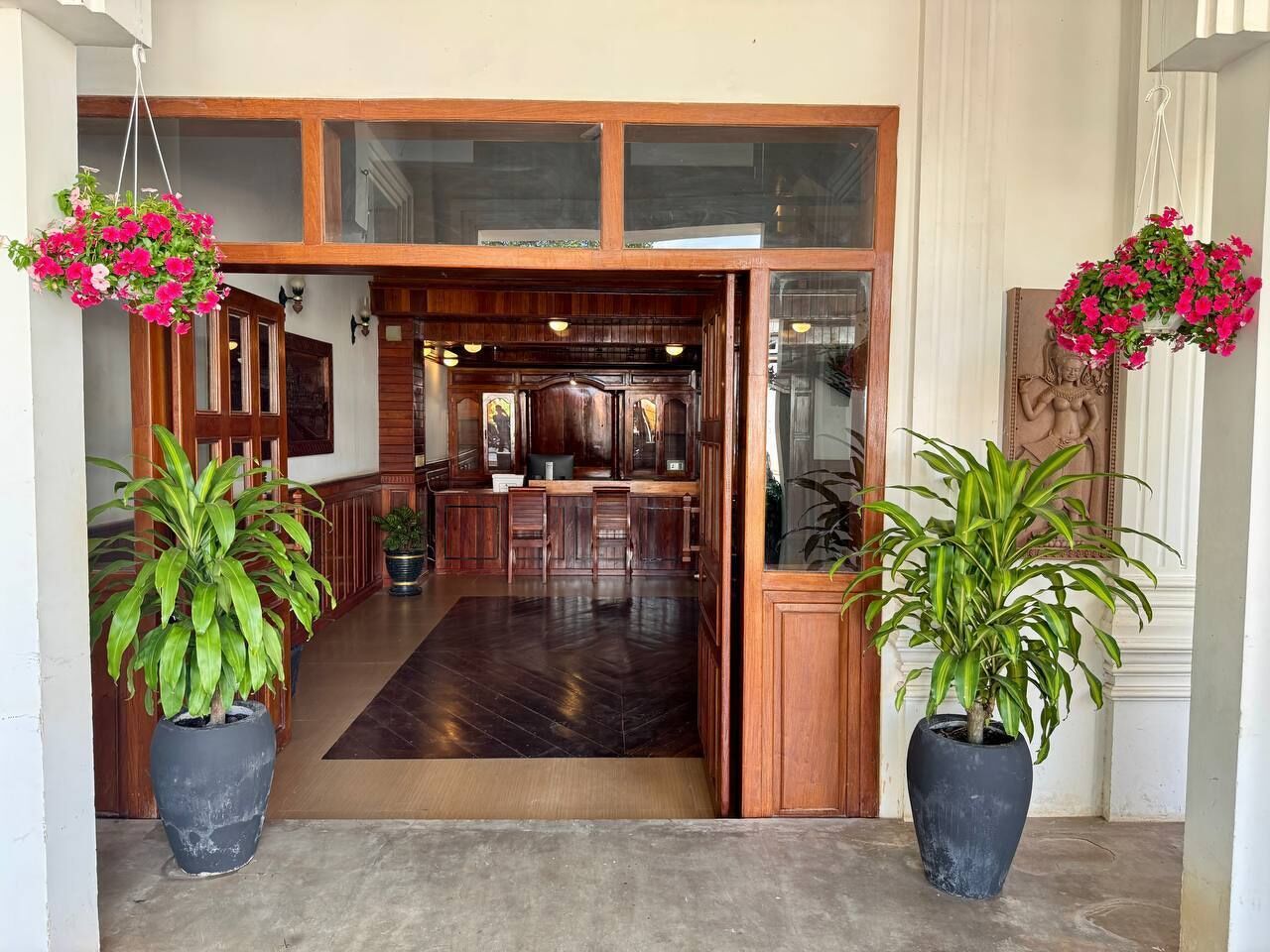 Un vestíbulo amplio con un mostrador de recepción de madera, dos grandes plantas en macetas y un suelo de madera oscura.