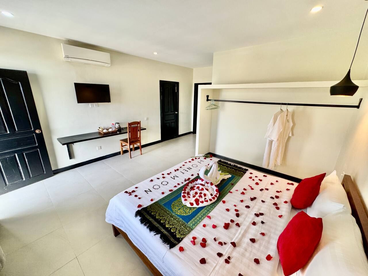 A hotel room with a bed adorned with rose petals and a towel, a television mounted on the wall, a desk with a chair, and a black door.