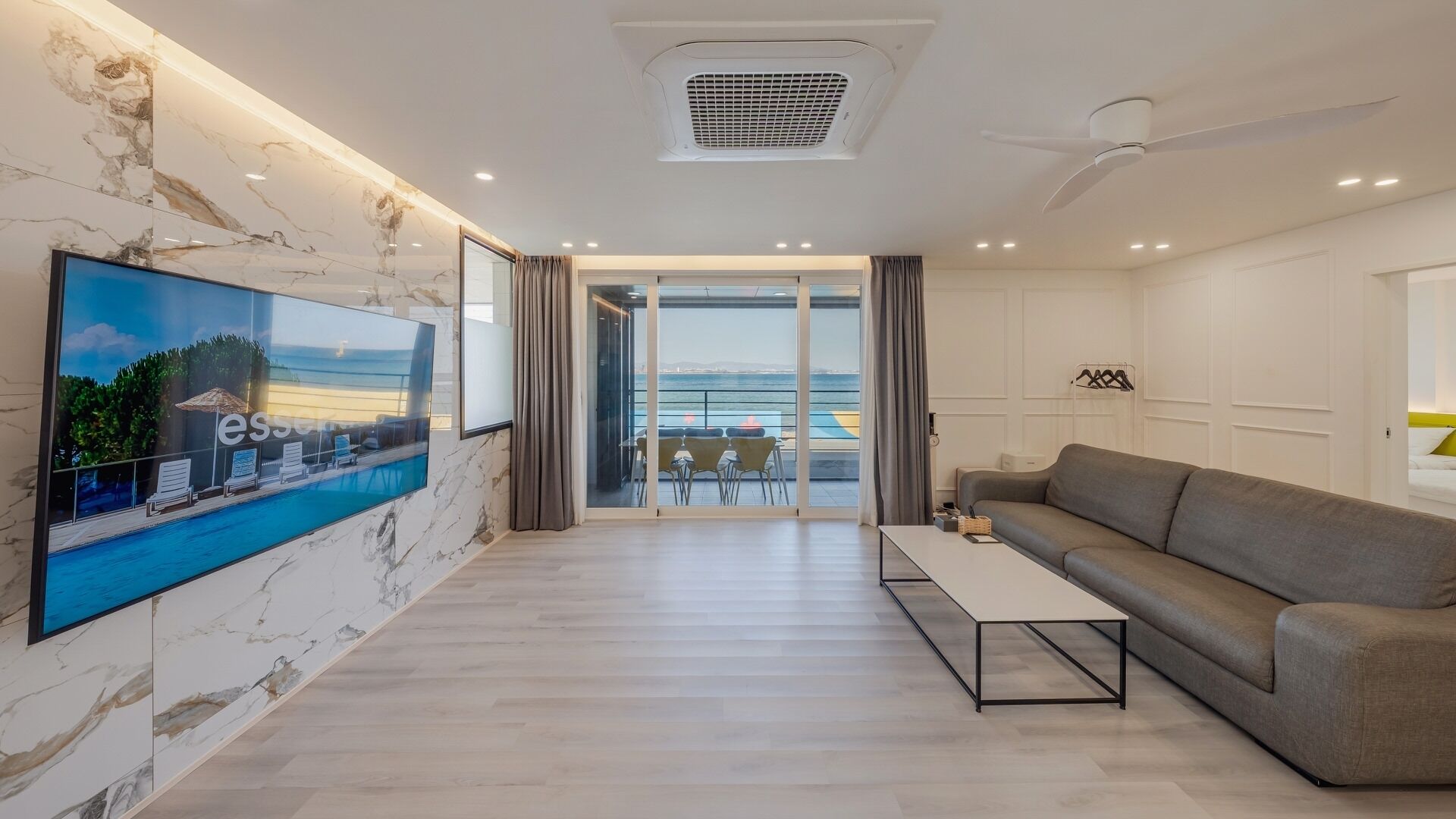 A modern living room with a large TV, a sofa, a coffee table, and a balcony with a view of the sea.