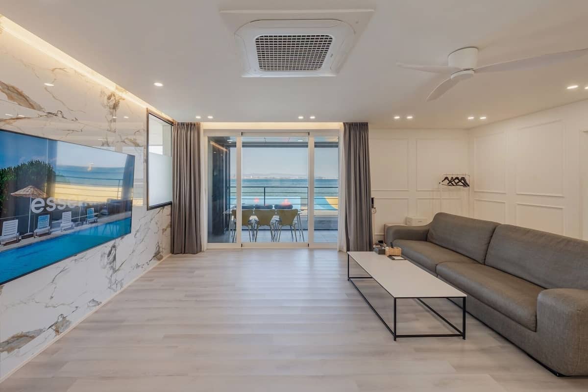 A modern living room with a large TV, a sofa, a coffee table, and a balcony with a view of the sea.