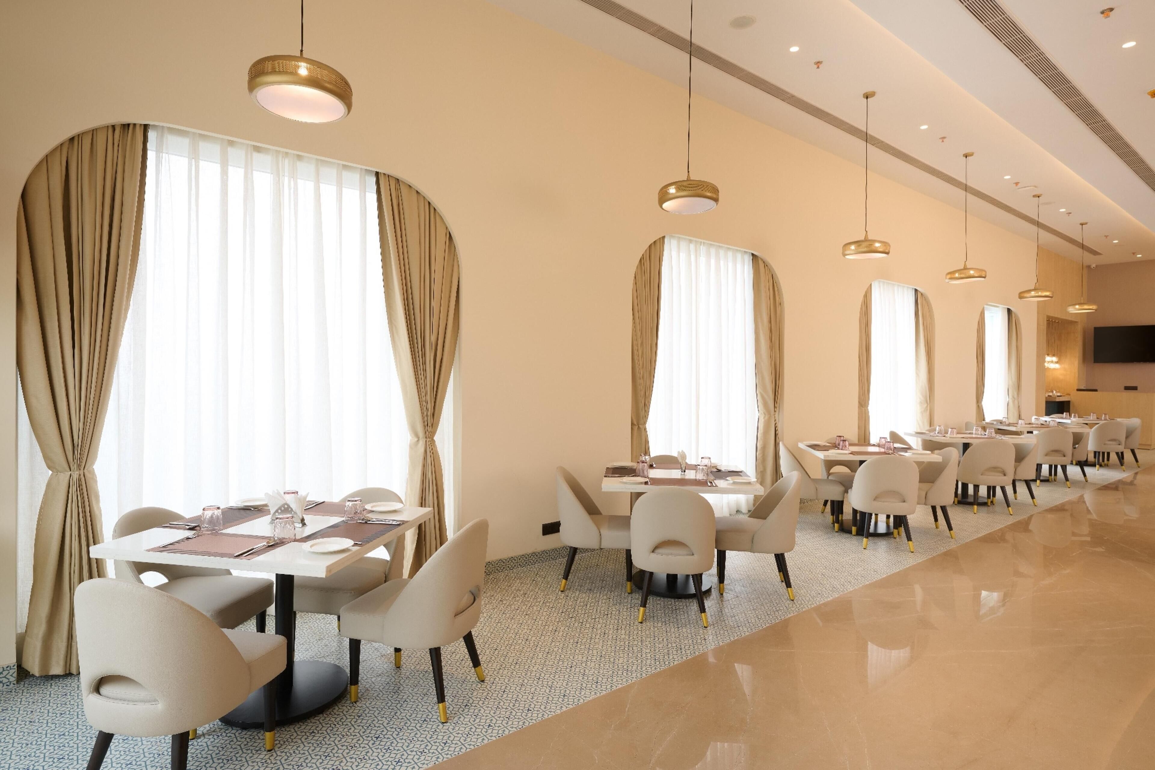 A hotel dining area with multiple tables and chairs, large windows with curtains, and pendant lighting.