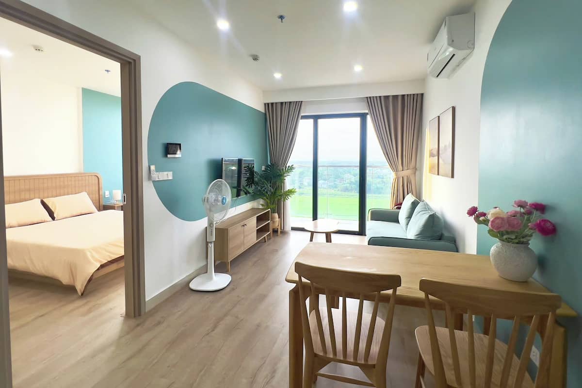 Une chambre d’hôtel comprenant un lit, un bureau avec des chaises, une télévision, un ventilateur et une grande fenêtre donnant sur un beau paysage.
