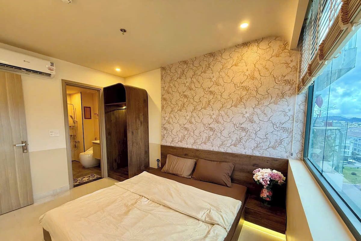 Une chambre avec un grand lit, une tête de lit en bois, une fenêtre donnant sur un beau paysage et une salle de bain visible à travers une porte ouverte.