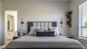 Une chambre moderne avec un grand lit, des tables de chevet, une étagère murale agrémentée de plantes et une fenêtre avec des stores.