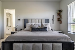 Ein modernes Schlafzimmer mit einem großen Bett, Nachttischen, einem Wandregal mit Pflanzen und einem Fenster mit Jalousien.