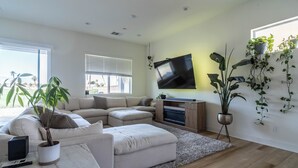 Un salon moderne avec un canapé d’angle, un téléviseur à écran plat fixé au mur et plusieurs plantes en pot.