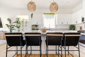 A modern kitchen with white cabinets, a central island with bar stools, hanging plants, and pendant lighting. - 3-Bedroom Pool House with Golf & Sunset Views (Indio)